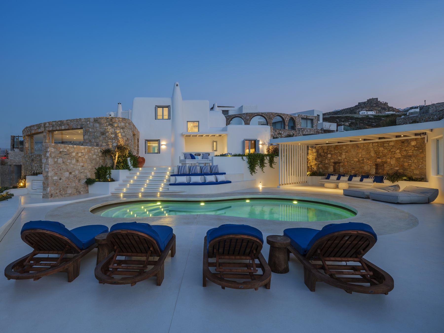 Villa KIYA at dusk: the lit pool, stone terraces and blue-cushioned loungers in front of the whitewashed house